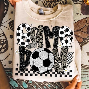 May include: A beige sweatshirt with the words "GAME DAY" in a mix of fonts and patterns. The word "GAME" features soccer ball and leopard print designs. A soccer ball graphic is centered below the text, with a checkered border.
