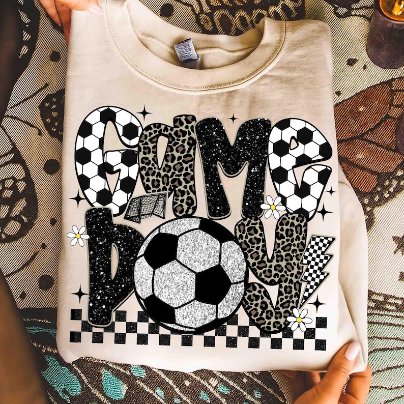 May include: A beige sweatshirt with the words "GAME DAY" in a mix of fonts and patterns. The word "GAME" features soccer ball and leopard print designs. A soccer ball graphic is centered below the text, with a checkered border.