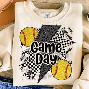 Op de afbeelding: Cr&egrave;mekleurige sweatshirt met een grafisch ontwerp met twee gele glitter softballs met rode stiksels, een bliksemschicht met een luipaardprint en twee sterren met een zwart-wit geblokt patroon. De woorden "Game Day" staan in het midden.