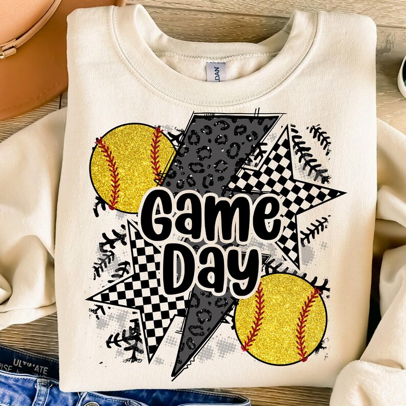 Op de afbeelding: Cr&egrave;mekleurige sweatshirt met een grafisch ontwerp met twee gele glitter softballs met rode stiksels, een bliksemschicht met een luipaardprint en twee sterren met een zwart-wit geblokt patroon. De woorden "Game Day" staan in het midden.