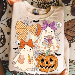 May include: Beige sweatshirt featuring a Halloween-themed graphic. The design includes cartoon ghosts with bows, books, and pencils, along with a floral pumpkin, spiderwebs, and bats. The bows have orange and purple patterns.