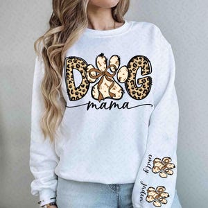 Puede incluir: Sudadera blanca con el texto "DOG mama" en una mezcla de estampados de leopardo y dise&ntilde;os de huellas de patas marrones. El dise&ntilde;o de la huella de la pata incluye un lazo y acentos de hueso. La manga presenta huellas de patas y los nombres "Emily" y "Jaden".