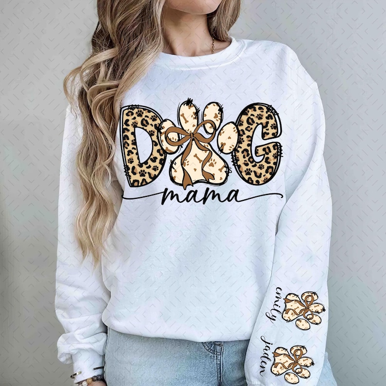 Puede incluir: Sudadera blanca con el texto "DOG mama" en una mezcla de estampados de leopardo y dise&ntilde;os de huellas de patas marrones. El dise&ntilde;o de la huella de la pata incluye un lazo y acentos de hueso. La manga presenta huellas de patas y los nombres "Emily" y "Jaden".