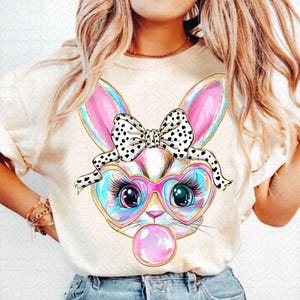 May include: A white t-shirt featuring a colourful cartoon bunny wearing heart-shaped glasses and a polka dot bow. The bunny is blowing a pink bubble.