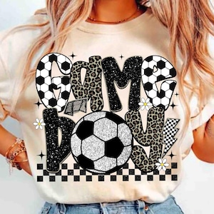 May include: A beige t-shirt with a soccer-themed graphic. The design features the words "GAME BOY" in a mix of fonts and patterns, including leopard print and soccer ball designs. A soccer ball is in the center.