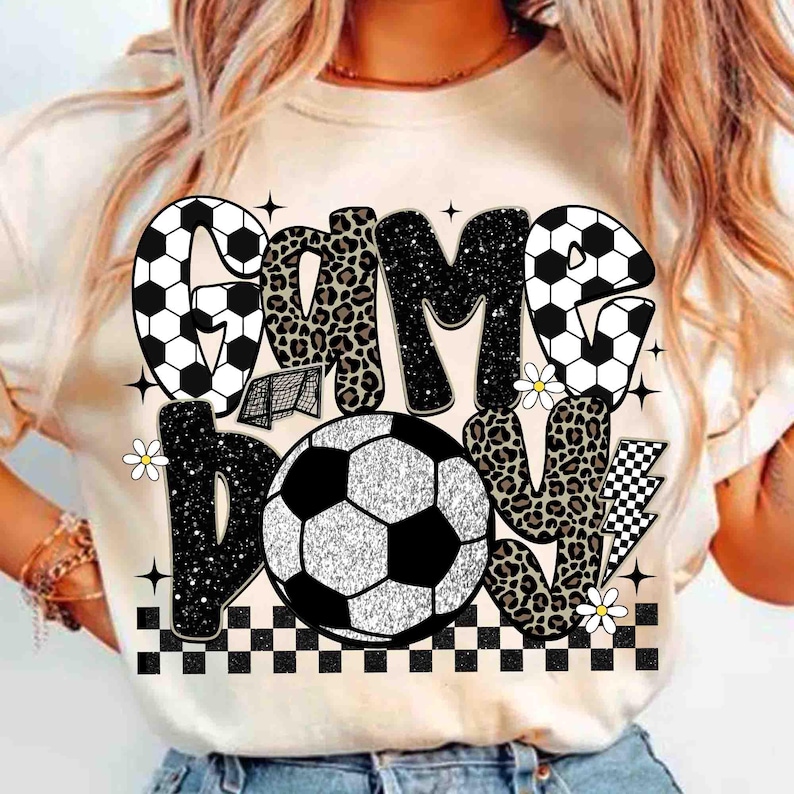 May include: A beige t-shirt with a soccer-themed graphic. The design features the words "GAME BOY" in a mix of fonts and patterns, including leopard print and soccer ball designs. A soccer ball is in the center.