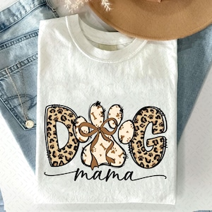 Puede incluir: Camiseta blanca con un dise&ntilde;o gr&aacute;fico con las palabras "DOG mama". Las letras "DOG" est&aacute;n en un estampado de leopardo, y la palabra "mama" est&aacute; en cursiva. Una huella de pata con un lazo est&aacute; en el centro.