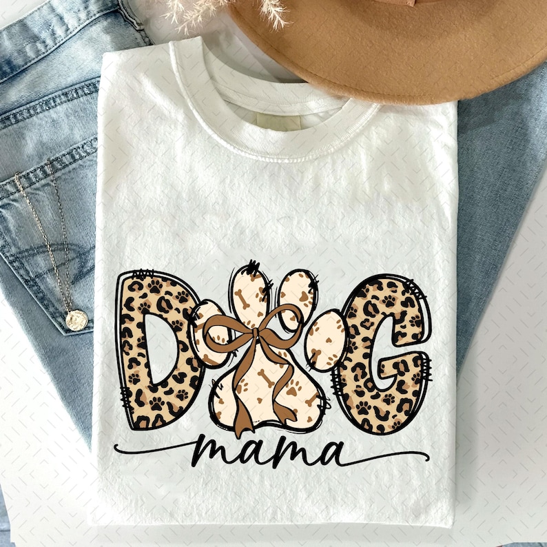 Puede incluir: Camiseta blanca con un dise&ntilde;o gr&aacute;fico con las palabras "DOG mama". Las letras "DOG" est&aacute;n en un estampado de leopardo, y la palabra "mama" est&aacute; en cursiva. Una huella de pata con un lazo est&aacute; en el centro.