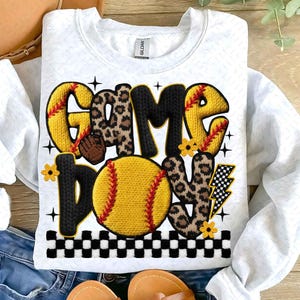 May include: White sweatshirt with a softball-themed design. The words "GAME DAY" are displayed in a mix of yellow, black, and leopard print, with a softball graphic. The design includes a baseball glove, flowers, and a lightning bolt.