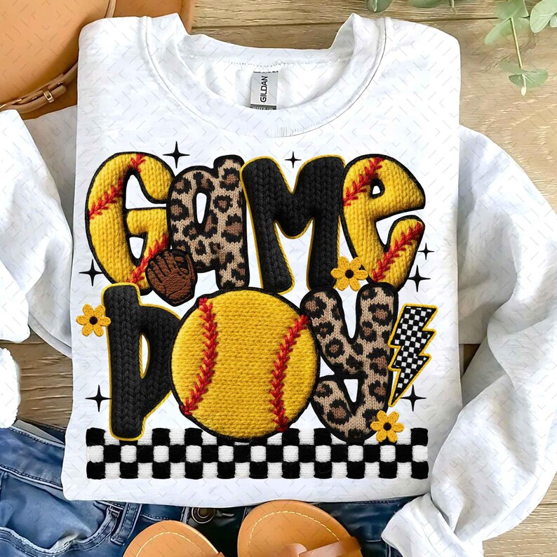 May include: White sweatshirt with a softball-themed design. The words "GAME DAY" are displayed in a mix of yellow, black, and leopard print, with a softball graphic. The design includes a baseball glove, flowers, and a lightning bolt.