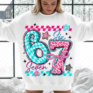 Könnte beinhalten: Weißes Sweatshirt mit einer großen Grafik mit den Zahlen "67" in einem lebendigen Design. Die Zahl "6" ist türkis und kariert, während die "7" rosa mit Leopardenmuster ist. Die Wörter "Six" und "Seven" sind ebenfalls enthalten.
