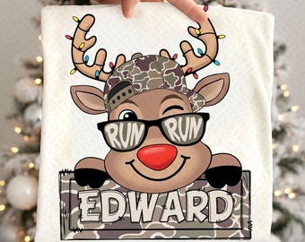 May include: A white t-shirt with a cartoon reindeer wearing a camouflage hat and sunglasses with "RUN RUN" on the lenses. The reindeer has a red nose and is holding a sign that says "EDWARD". The antlers are decorated with Christmas lights.