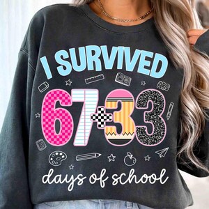 May include: Dark gray sweatshirt with the text "I SURVIVED 67+33 days of school" in light blue and white. The numbers are decorated with pink, black, and yellow patterns, and school-themed graphics.