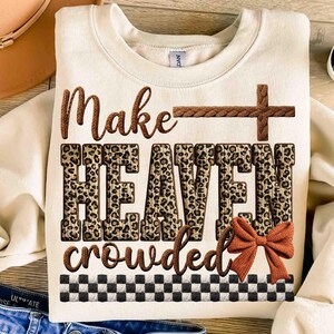 May include: Cream-colored sweatshirt with the phrase "Make HEAVEN crowded" in brown and leopard print. A brown cross and bow accent the design. A black and white checkered pattern is at the bottom.