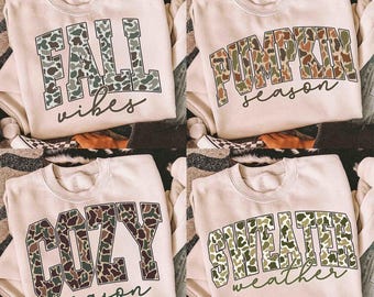 May include: Four cream-colored sweatshirts with fall-themed text. The sweatshirts feature the phrases "FALL vibes", "PUMPKIN season", "COZY season", and "SWEATER weather" in a camouflage pattern. The text is in a bold, collegiate-style font.