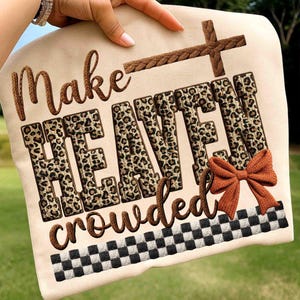 May include: A beige fabric item with the words "Make HEAVEN crowded" in brown script and leopard print letters. A brown cross and bow accent the design, with a black and white checkered border.