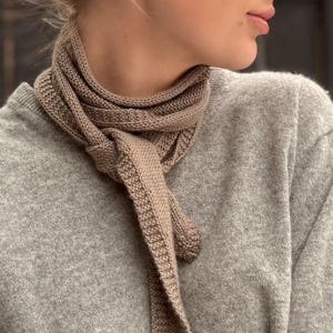 Könnte beinhalten: Nahaufnahme eines gestrickten Schals in Hellbraun, um den Hals gebunden. Der Schal besteht aus einem weichen, strukturierten Material und wird über einem grauen Pullover getragen. Die Enden des Schals hängen herunter und erzeugen einen stilvollen Look.