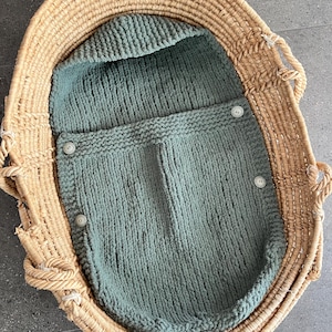 Puede incluir: Un saco de dormir para bebé de punto verde salvia con capucha y cierres de botón, dentro de una cesta tejida.
