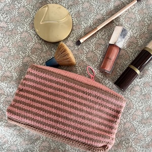 May include: A collection of makeup items, including a gold compact, makeup brushes, lip gloss, and a lipstick. A pink and brown striped knitted cosmetic bag is in the foreground. The items are arranged on a floral patterned fabric.