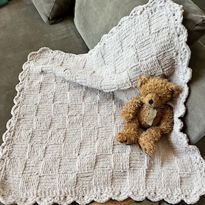 May include: A light grey, woven baby blanket with a scalloped edge and a brown teddy bear with a black and white striped bow. The blanket has a textured, basketweave pattern.