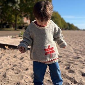 May include: A light gray long-sleeve knit sweater with a ribbed neckline and cuffs, featuring a red embroidered graphic of a stylized tree on the front. The sweater is paired with blue denim jeans and gray slip-on shoes, creating a casual outfit.