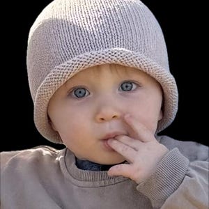 Può includere: Un bambino con occhi azzurri brillanti indossa un cappello di maglia beige chiaro e una maglietta a maniche lunghe abbinata. Il bambino ha un dito in bocca, suggerendo un momento di contemplazione.