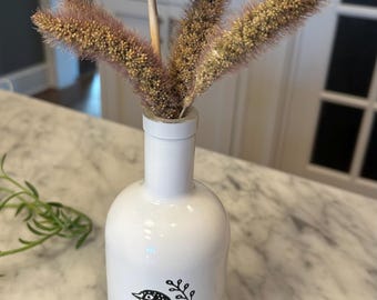 May include: A white ceramic vase with a black bird illustration and dried floral arrangement. The vase holds a large, brown, layered flower and several sprigs of tan and purple grasses. The vase is on a marble surface.