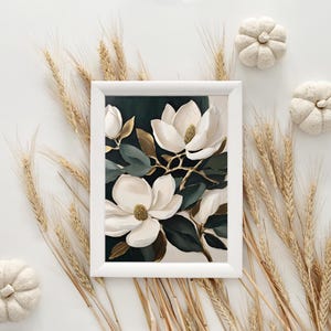 K&ouml;nnte beinhalten: Wei&szlig; gerahmter Kunstdruck mit wei&szlig;en Magnolienbl&uuml;ten mit goldenen Akzenten und dunkelgr&uuml;nen Bl&auml;ttern. Das Bild wird durch Weizen und kleine wei&szlig;e K&uuml;rbisse erg&auml;nzt, was eine herbstliche &Auml;sthetik hervorruft.