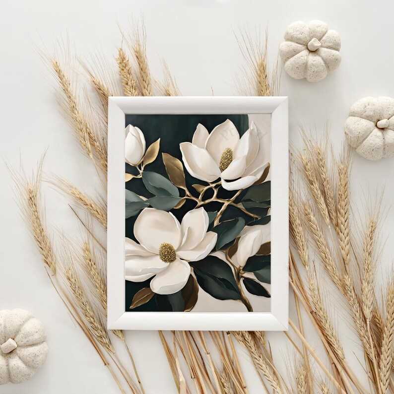 K&ouml;nnte beinhalten: Wei&szlig; gerahmter Kunstdruck mit wei&szlig;en Magnolienbl&uuml;ten mit goldenen Akzenten und dunkelgr&uuml;nen Bl&auml;ttern. Das Bild wird durch Weizen und kleine wei&szlig;e K&uuml;rbisse erg&auml;nzt, was eine herbstliche &Auml;sthetik hervorruft.