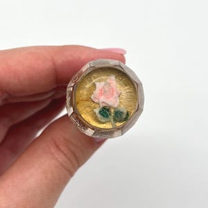 May include: A decorative bottle stopper with a faceted, clear glass body and a gold-coloured interior. The stopper features a miniature pink rose with green leaves. The stopper is held in a hand against a white background.