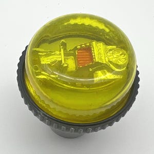 May include: A yellow, round bottle stopper with a black base. The stopper has a detailed design inside the yellow dome, featuring a building with a red roof. The base has a ridged design.