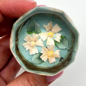 May include: A small, round, light blue trinket box with a floral design. The interior features a light blue base with three white and yellow flowers and green leaves. The box has a decorative, textured edge.