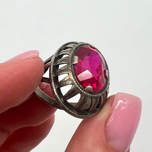 Pode incluir: Um anel prateado com uma grande pedra preciosa rosa brilhante em forma oval. O anel tem um design vazado em torno da pedra preciosa, com uma configuração detalhada de inspiração vintage. O anel é mostrado em um fundo branco liso.