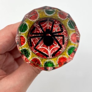 Puede incluir: Un tapón de botella decorativo con un diseño facetado. El tapón presenta un patrón rojo, verde y dorado alrededor del borde, con un diseño de araña negra en el centro. El tapón se sostiene en una mano sobre un fondo blanco.
