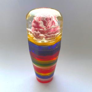 May include: A colorful, decorative shift knob. The top is clear with a pink flower inside. The body is a layered design of yellow, blue, red, green, and other colors. The knob is cylindrical and stands upright.