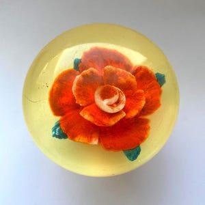 May include: A clear, yellow paperweight with a vibrant orange rose encased inside. The rose has detailed petals and green leaves, creating a three-dimensional effect. The paperweight is round and sits on a white surface.