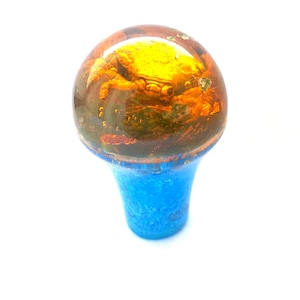 Op de afbeelding: Een decoratieve kastknop met een ronde, amberkleurige bovenkant en een blauwe basis. De amberkleurige bovenkant heeft een getextureerd, gespikkeld uiterlijk met hints van goud en bruin. De basis is een levendige, effen blauwe kleur.