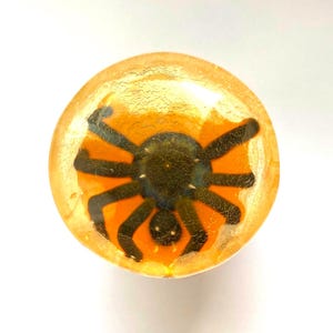 May include: A round, orange-yellow object with a black spider design. The spider has eight legs and a dark body, set against an orange background. The object has a textured, cracked appearance, suggesting a resin or glass material. The design is centered, creating a Halloween theme.