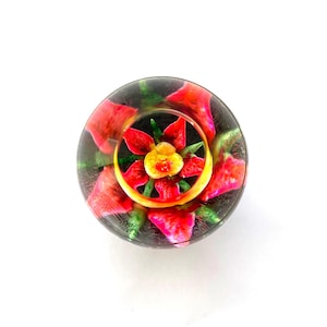 Puede incluir: Un tapón de botella de vidrio transparente con un diseño floral. El tapón presenta una flor amarilla central rodeada de pétalos rojos y hojas verdes, sobre un fondo oscuro. El diseño está encerrado en vidrio transparente.