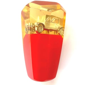 May include: A decorative car-themed object. The top portion is a transparent amber color, encasing a gold-colored vintage car model. The bottom section is a solid red color, with a faceted design. The object is set against a white background.