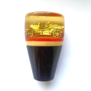 May include: A vintage-style car gear shift knob. The top section is clear with a gold-colored car design. Below the clear section is a red and cream-colored band, and the base is dark brown.