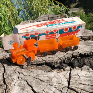 May include: Orange plastic toy train with three cars. The train has a black chimney and wheels. The box is white with red and blue accents and the text "ЗАВОД ИГРУШКА" in Cyrillic.