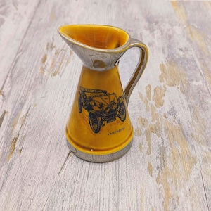 Puede incluir: Jarra pequeña de cerámica vintage con acabado amarillo y plateado. La jarra presenta una ilustración negra de un coche antiguo y la palabra "Lancaster". El asa y el pico son plateados, y el cuerpo es amarillo.