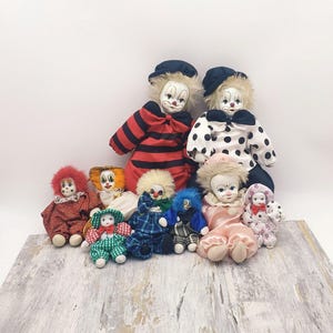 May include: A collection of nine porcelain clown dolls. The dolls are all dressed in colorful costumes with various patterns, including stripes, polka dots, and plaid. Some dolls have red, white, and blue accents.