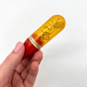 May include: A vintage toy car inside a clear, yellow, capsule-shaped container. The base is red, with a silver band separating it from the yellow top. The toy car inside is a detailed miniature.