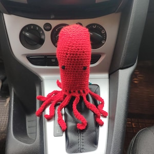 May include: A bright red, crocheted octopus shift knob cover. The octopus has a rounded head, small black eyes, and multiple tentacles. It is installed on the gear shift of a car, adding a whimsical touch to the interior.