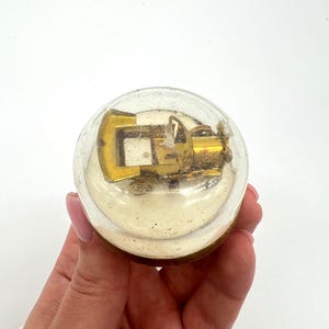 May include: A clear, round glass ornament containing a miniature gold-coloured tractor. The ornament is held in a hand against a white background. The tractor has a detailed design.