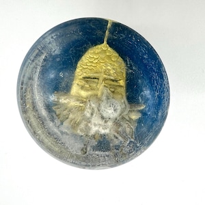 May include: A round, blue candle with a gold-colored, ornate design in the center. The design appears to be a stylized face or mask. The candle has a textured, almost frosted appearance.