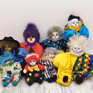 以下が含まれることがあります： ペイントされた顔とカラフルな服を着た、ヴィンテージのピエロ人形のコレクション。人形は、さまざまな髪型、帽子、水玉模様やチェック柄などのパターンを特徴としています。蝶ネクタイやフリルカラーのものもあります。人形は明るい色の木製の表面に配置されています。