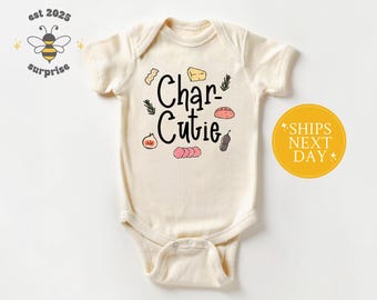 May include: Cream-colored baby onesie with the text "Char-Cutie" and food illustrations. The design includes a bee logo and the text "est 2025 surprise". The onesie has short sleeves and a snap closure.